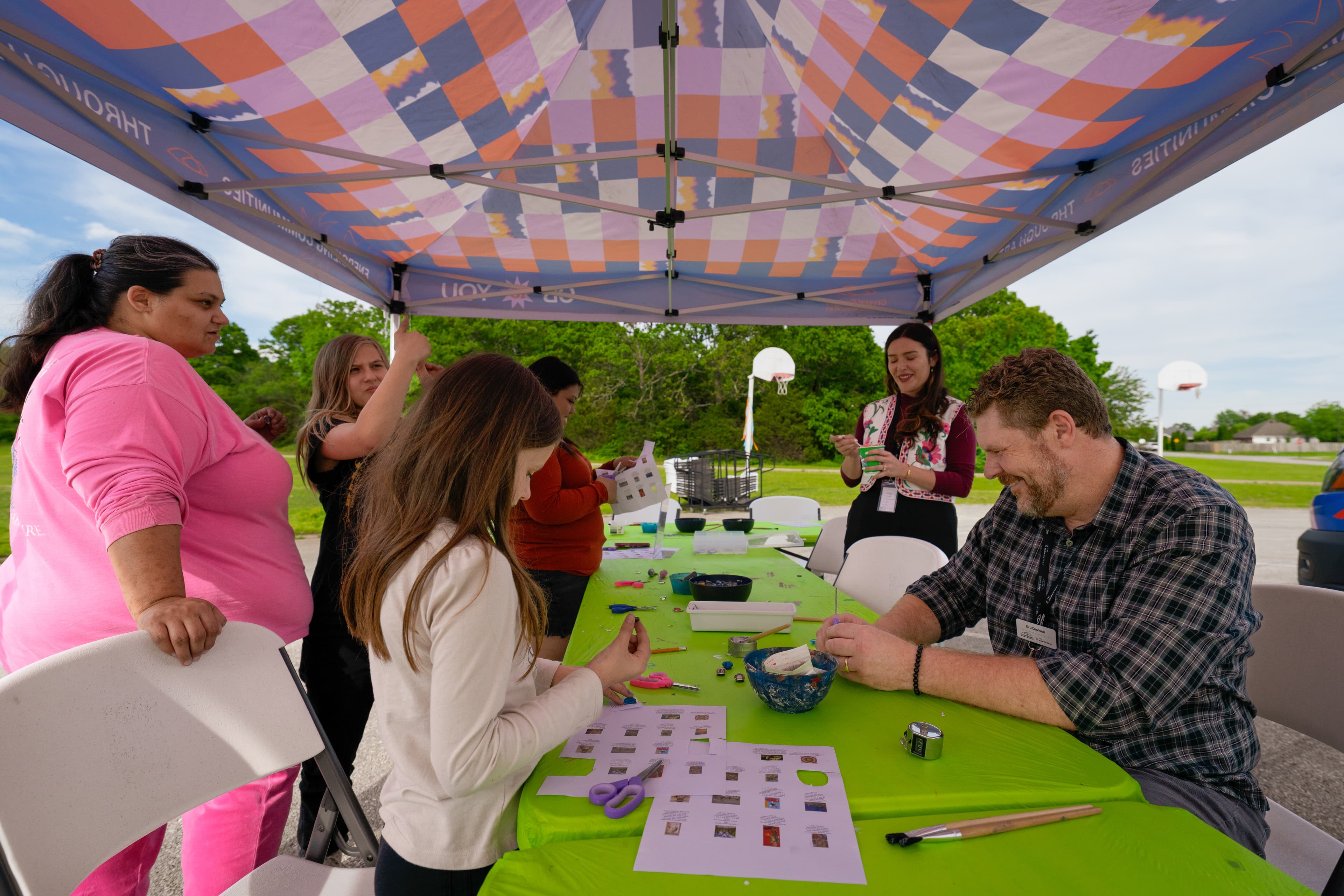 Children and adults gathered around a bright green table creating art outdoors under a colorful canopy.